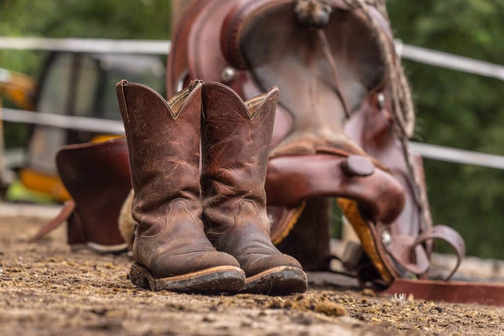 The Trainer's Loft Riding Boots, Ranch life scenery: muddy western boots in front of a western saddle. Cowboy boots. Muddy working boots