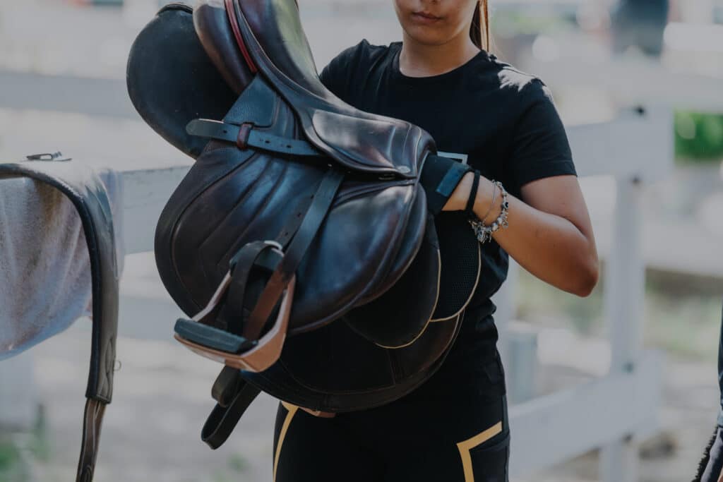 The Trainer's Loft Leather Care, Close-up of a woman holding a horse saddle at an outdoor stable, ready for equestrian activities. Captures the essence of preparation and care in horseback riding.